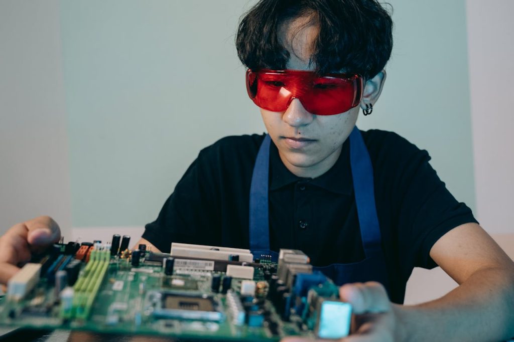 A young technician with red goggles examines a computer circuit board in a workshop setting.